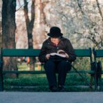 person sits on bench reading book in front of tree at daytime